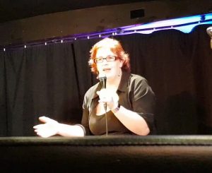 image of kitty fitton, a woman with red hair in a black outfit talking into a microphone with one arm outstretched. 
