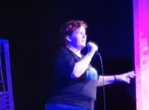 Kitty Fitton holding a microphone in her right hand talking on stage. 
