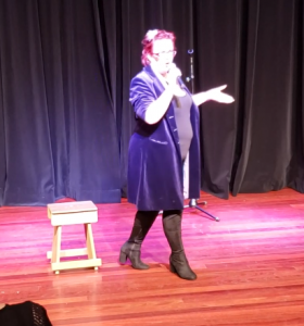 Kitty Fitton wearing a long purple coat and tall boots walking across the floor talking into a microphone. 