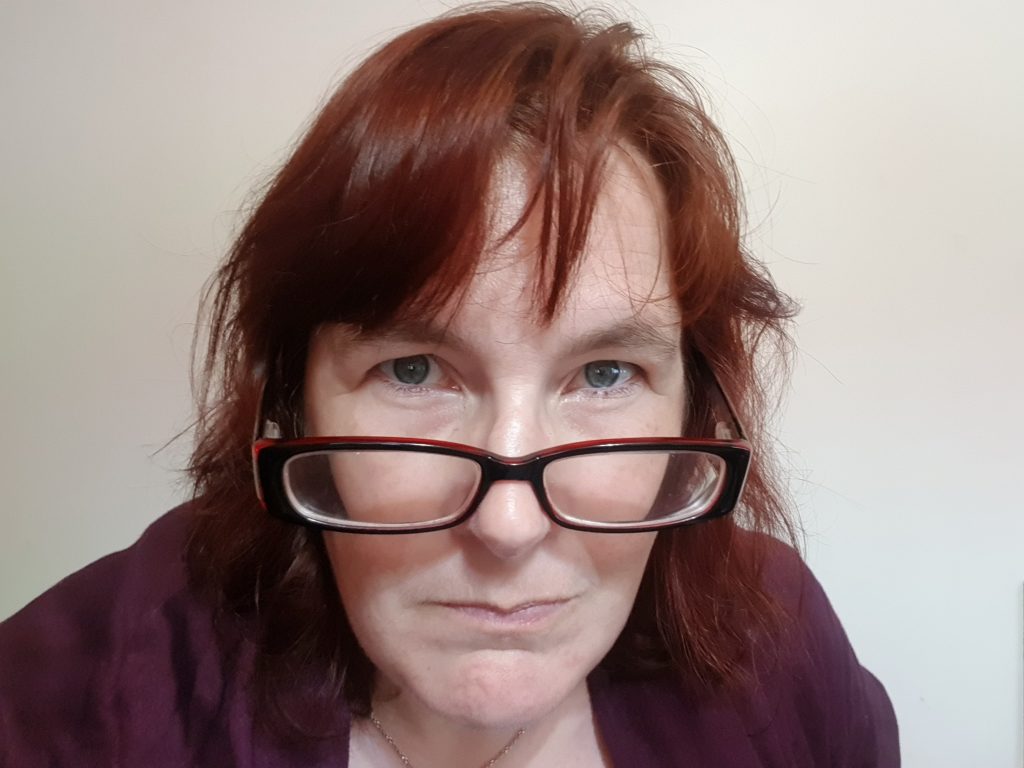 woman with red hair and plum shawl looking directly into the camera over red spectacles.