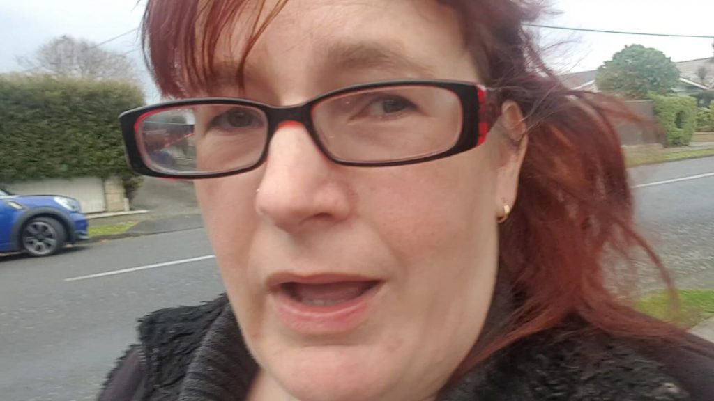 woman looking into the camera wearing glasses. She is outside in a street with a road in the background.