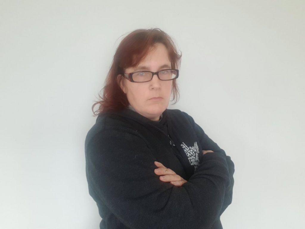 red haired woman in grey hoodie and glasses looking crossly into the camera with folded arms.