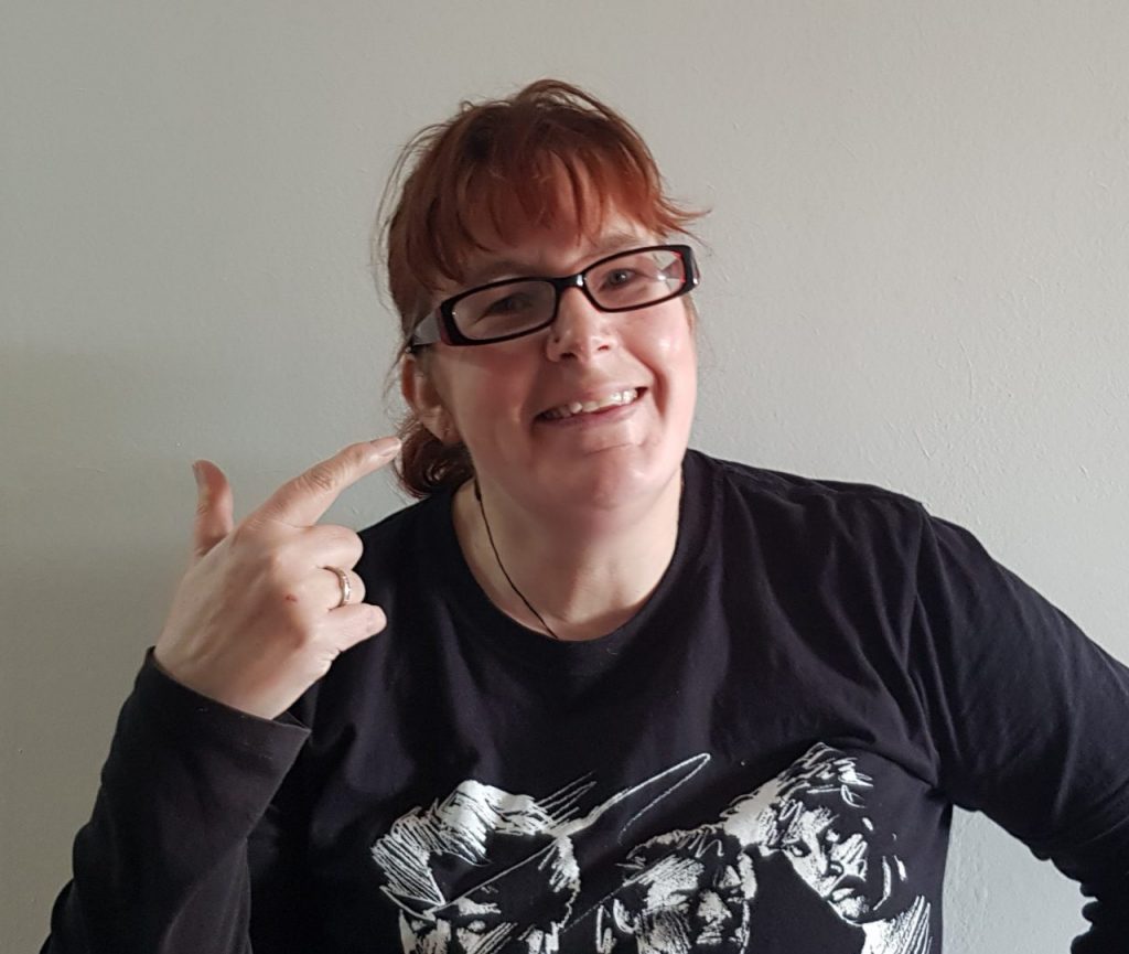 kitty fitton with red hair and glasses pinting aat herself and grinning.