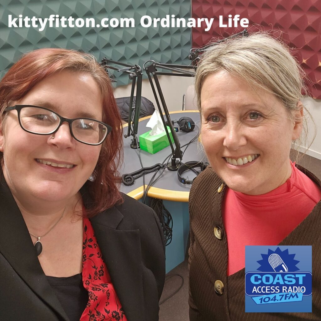 Kitty Fitton with red hair on the left and Catherine Scullen with blonde hair on the right smile into the camera with a recording studio in the background.
