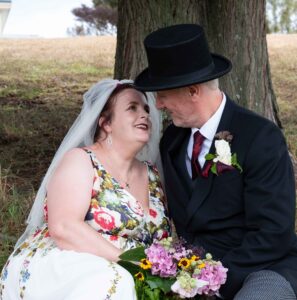 image of kitty in a wedding dress and mike in top hat and tailsuit sat under a tree gazing happily at each other.