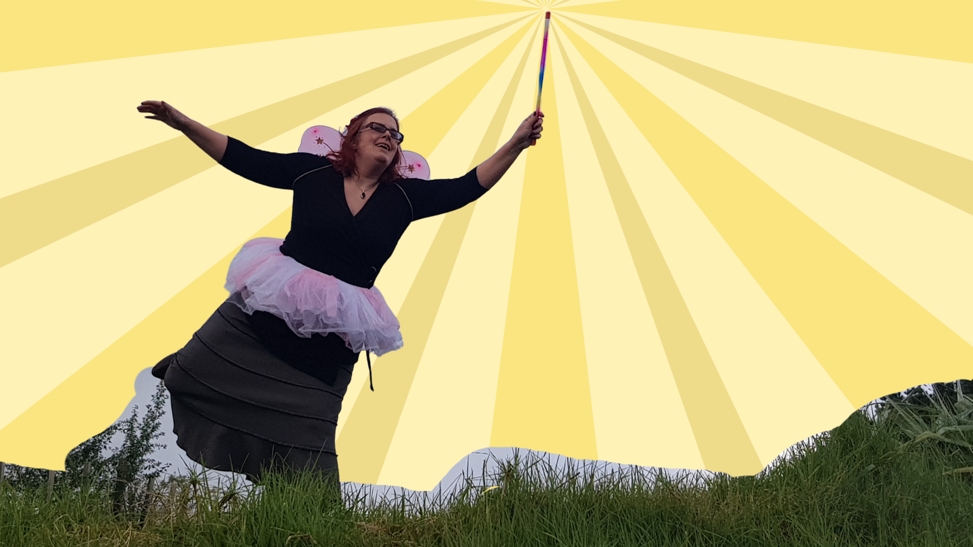kitty doyle in a fairy tutu over a long brown skirt with pink fairy wings and a wand balancing on  one leg grinning with arms outstretched. 