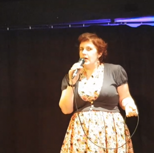 image of kitty fitton holding a microphone in a bright yellow and pink dress. 